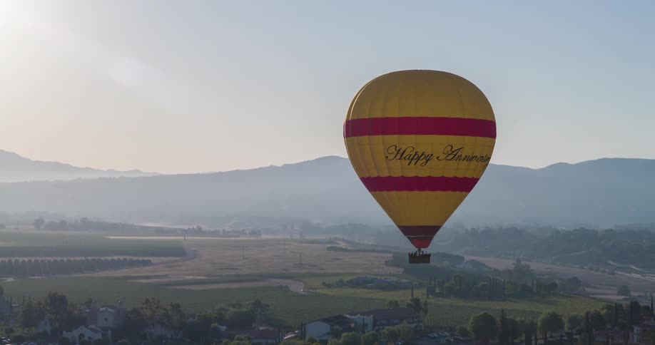 A yellow and red hot air balloon with happy anniversary written on it floats above a sunlit valley