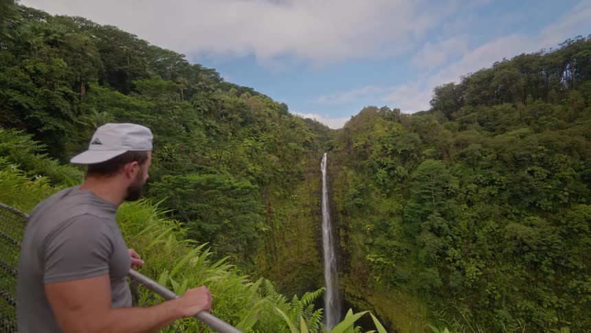 The video shows a powerful waterfall cascading down a sheer rock face into a pool below surrounded by a dense lush green jungle the camera captures the scene from a high angle looking down at the waterfall