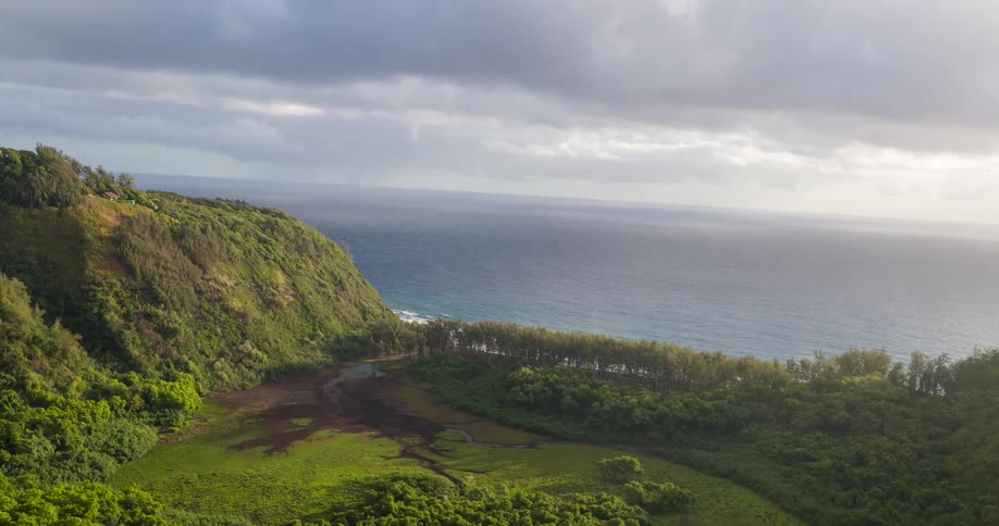 A steep lush green hillside next to the ocean with white waves crashing on the shore below a sky filled with gray and white clouds