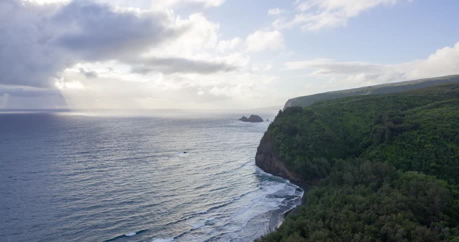 The video shows a scenic view of a coastline with a steep green cliff face meeting the ocean waves crash against the shore below the cliff and a dramatic cloudy sky is visible with a beam of sunlight breaking through