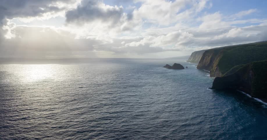 A sweeping aerial view shows sunlight breaking through the clouds to illuminate the vast ocean along a dramatic shadowy coastline