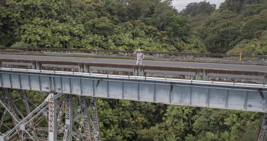 The video shows a man standing on an old metal bridge that spans a lush green valley the man wearing a white hat and a gray tshirt turns and walks to the right side of the screen
