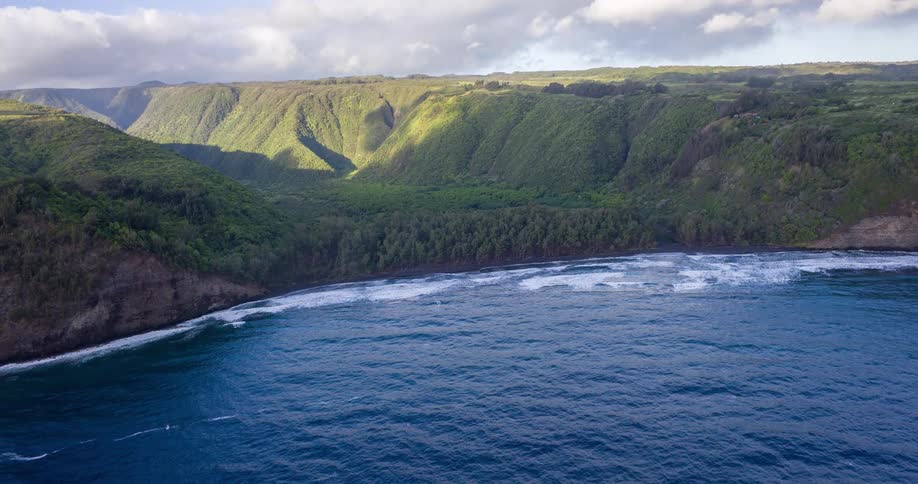 A mountainous island covered in green vegetation at midday as ocean waves hit the rocky shoreline