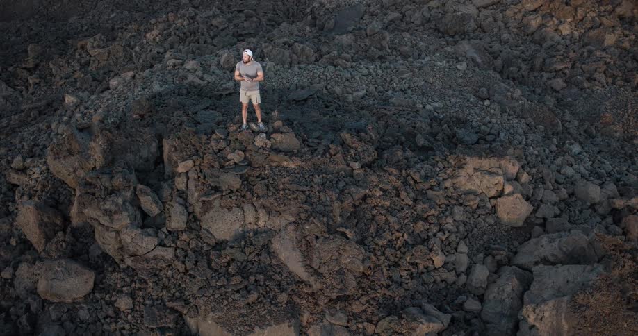 A man wearing a white backward baseball hat shorts sneakers and an ash tshirt stands on the peak of a rock in a rocky terrain at midday
