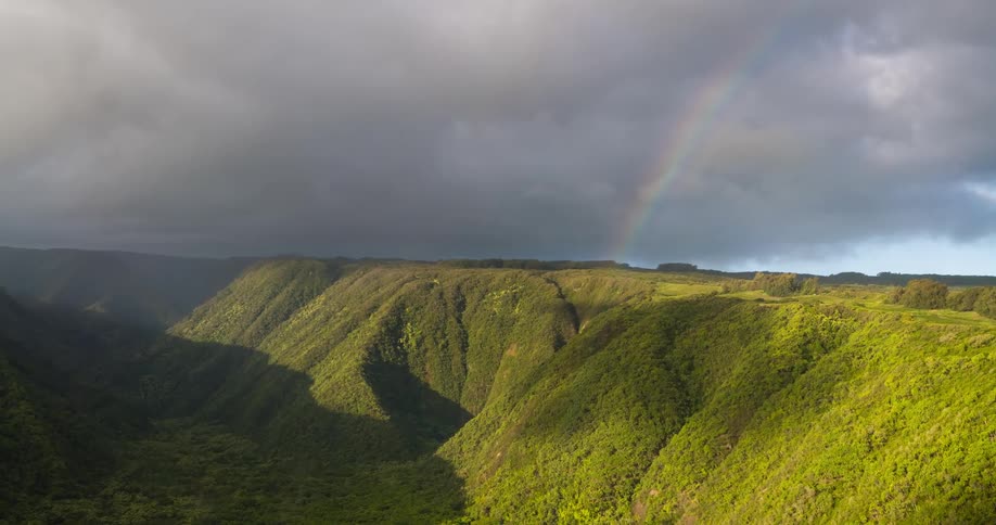 A rainbow emerges from stormy clouds over a majestic sunlit green valley
