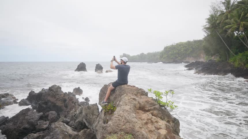 The video shows a man sitting on a rocky outcrop by the ocean taking a picture with his phone the ocean is choppy and there are large rock formations in the water with palm trees and other foliage visible on the shore in the background