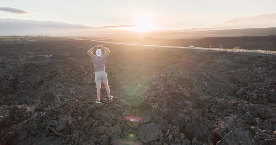 A man stands triumphantly with arms raised on a vast volcanic lava field as the sun sets over the distant mountains and coastline