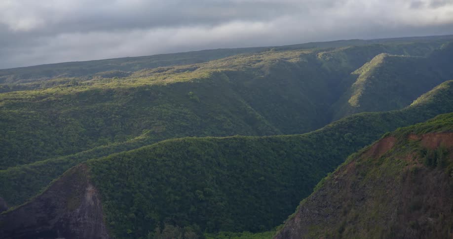 A wide terrain of mountains and hills covered in green vegetation at midday with dark clouds covering the sky