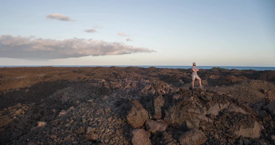 A man in shorts and a cap stands confidently on rugged volcanic rock with the ocean and clouds in the distance