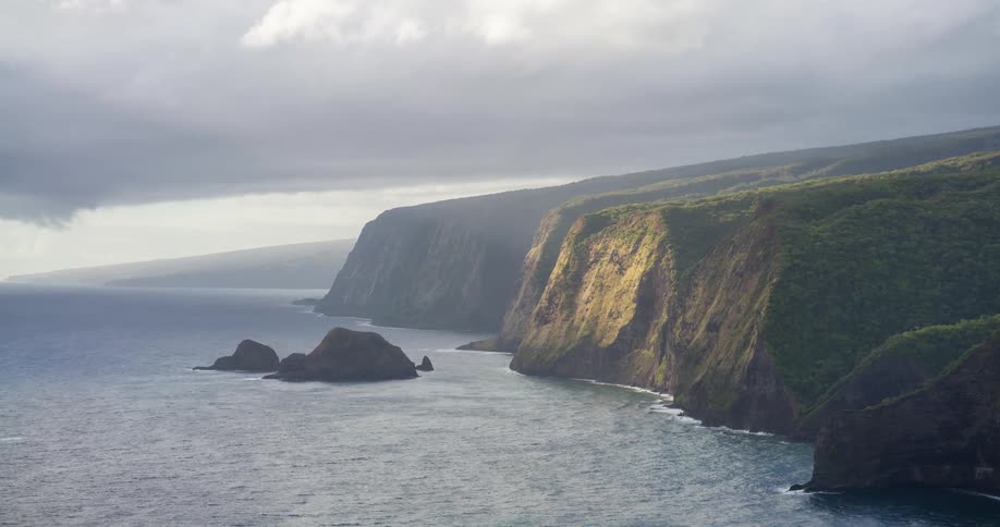 A mountainous island on a cloudy day with ocean waves hitting the rocky shore