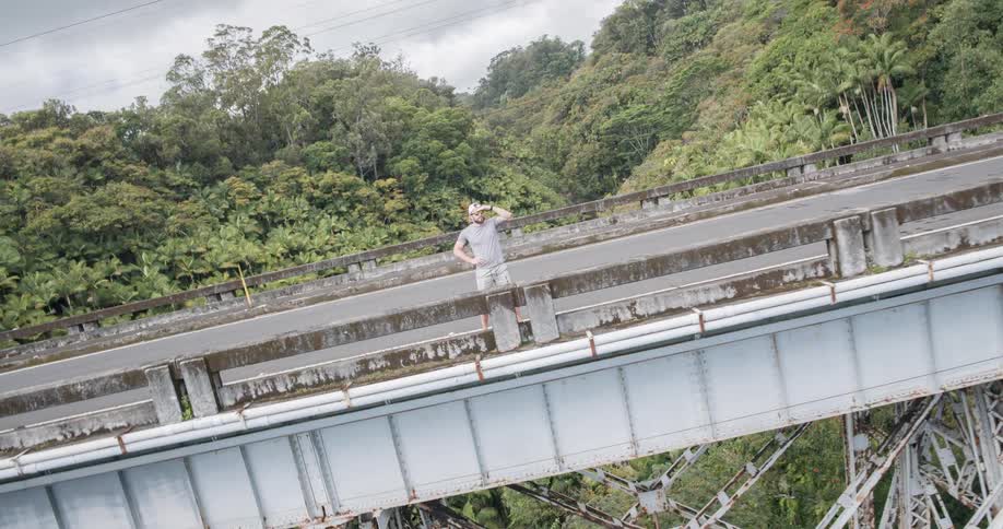 A man standing on a bridge wearing a hat staring into the distance with the bridge surrounded by forest trees at midday