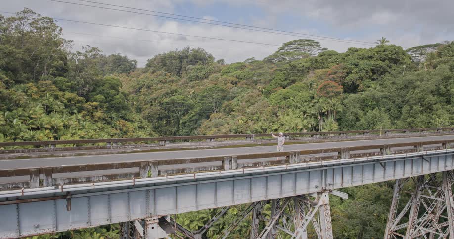 A tall metal viaduct spans a deep valley covered in lush green trees with a man wearing a white tshirt and shorts standing in the middle of the empty bridge filming with a phone
