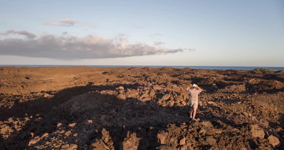 The video shows an aerial shot of a man standing on a rocky desolate landscape which appears to be a lava field he is looking out at the ocean in the distance as he adjusts his hat