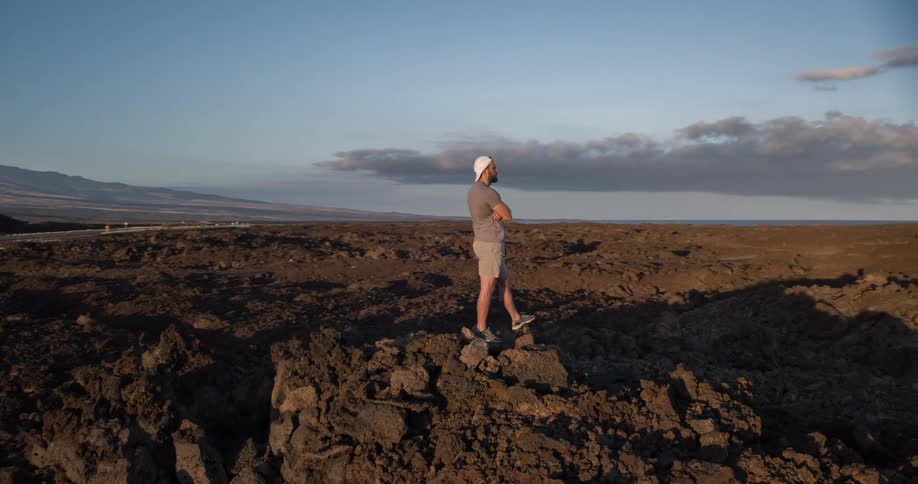 A man stands with his arms crossed on a vast volcanic lava field looking out at distant mountains and the sea