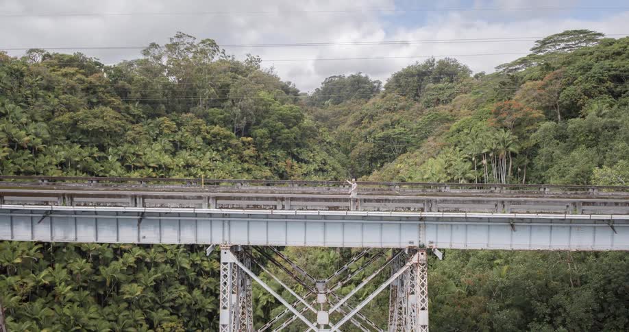 A man wearing a hat and shorts walks on a bridge recording a selfie video at midday with the bridge surrounded by trees