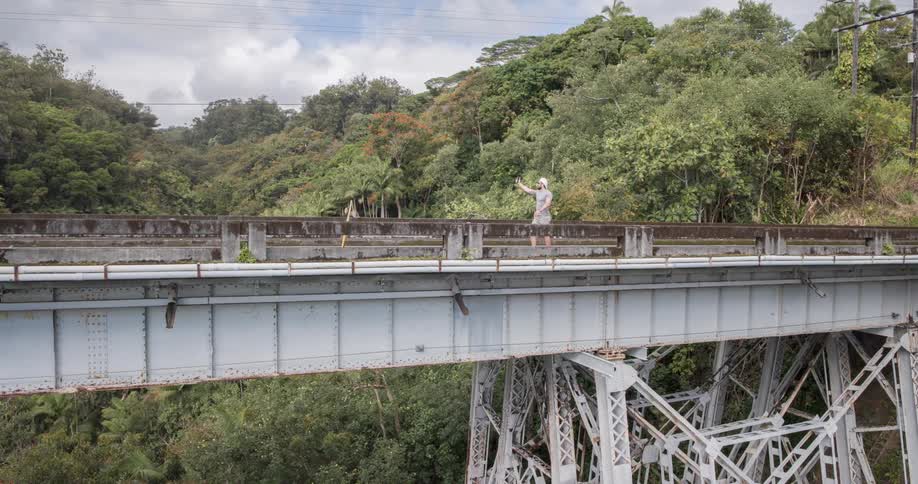 A man wearing a hat and shorts walks on a bridge recording a selfie video as a car passes by at midday with the bridge surrounded by trees