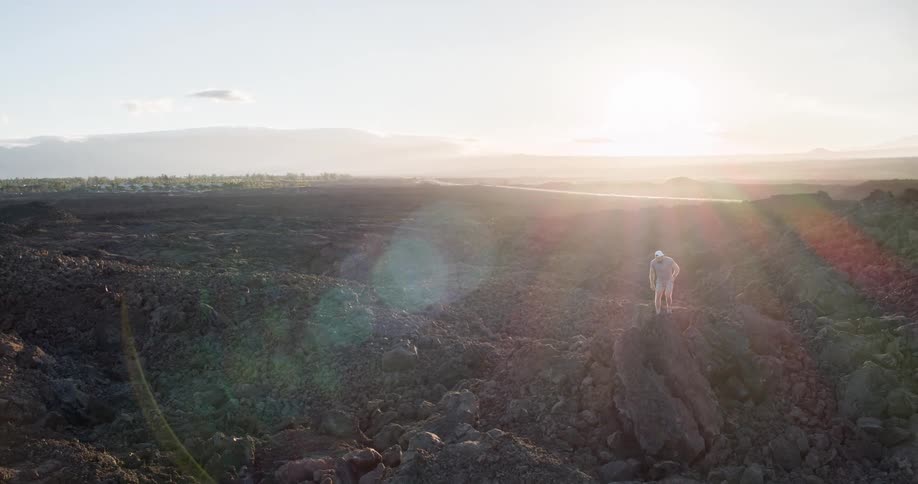 The video shows a wide aerial view of a desolate rocky landscape likely a lava field a person is seen walking carefully down a large rock formation in the foreground with mountains visible in the hazy distance
