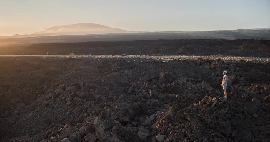 The video shows an aerial shot of a man standing on a rocky dark landscape possibly a lava field he is looking out at the horizon where the sun is setting with mountains in the distance