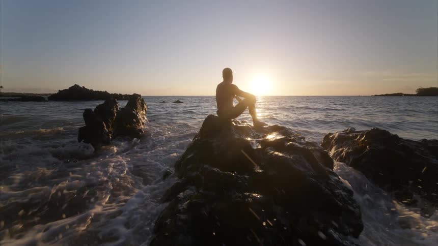 Man sitting on rocks along the coast as waves roll in and splash around him