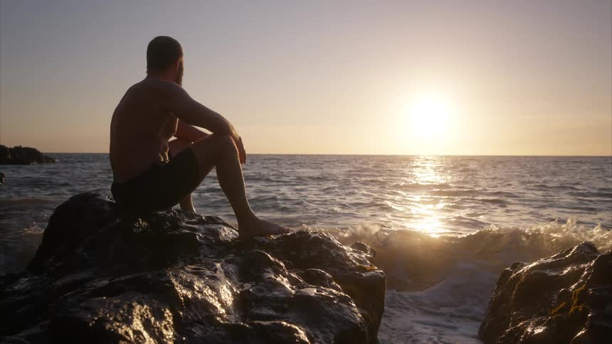 A man shirtless and barefoot sit on a rock on shore as the ocean waves crash agressively on the rock with the orange sun setting in a distance