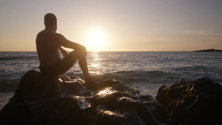 A man sits on a rocky coastline watching the sunset as ocean waves crash against the rocks