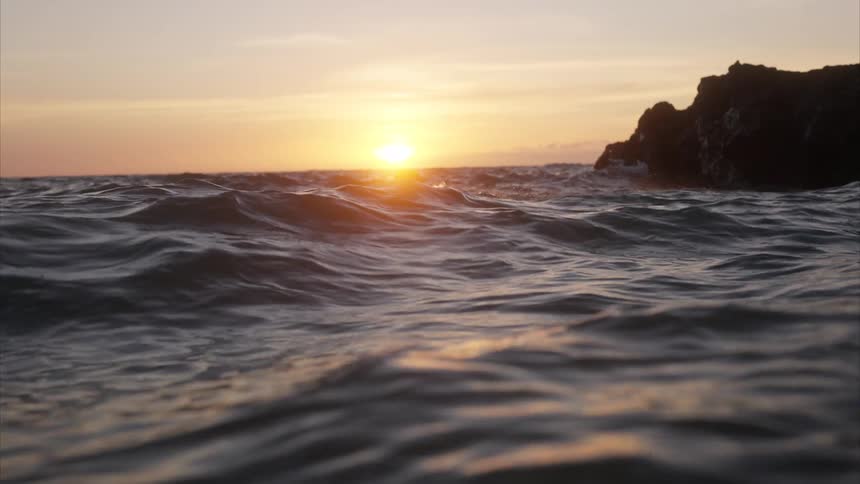 A swimmer is performing the freestyle stroke in the ocean at sunset with a rocky cliff in the background as the sun casts a bright reflection on the water