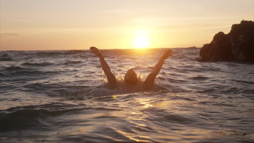 Man swims in shallow water off the coast as the sun glows on the horizon