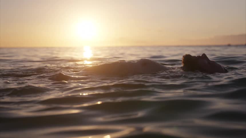 A person floats peacefully on their back in the calm ocean silhouetted by the golden glow of a beautiful sunset
