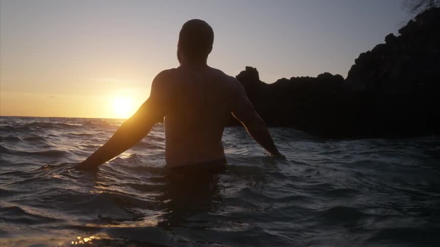 Man wearing swim trunks stands in the water near the shore while the sun sits on the red tinted horizon