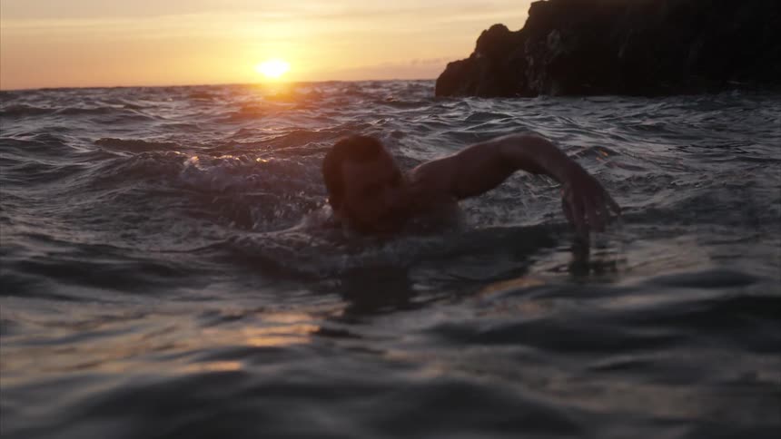 Man swimming in the ocean near the coast turns to face the sun sitting on the horizon