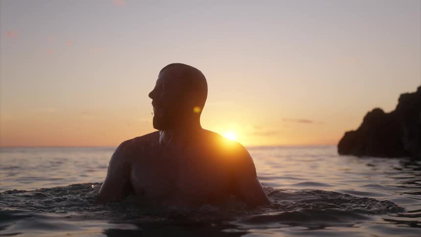 A man emerges from the ocean and shakes off water from his face silhouetted against the beautiful golden sunset