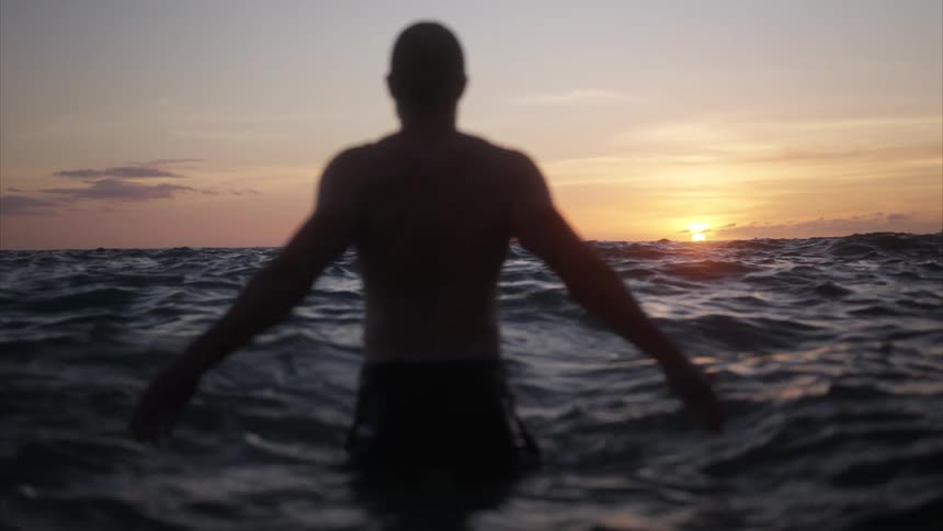 A man stands waistdeep in the ocean water enjoying the moment as ripples form from the waves with the orange setting sun in the distance