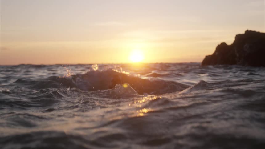 A man swims in the ocean backstroking farther out with the setting sun and a large rock behind him
