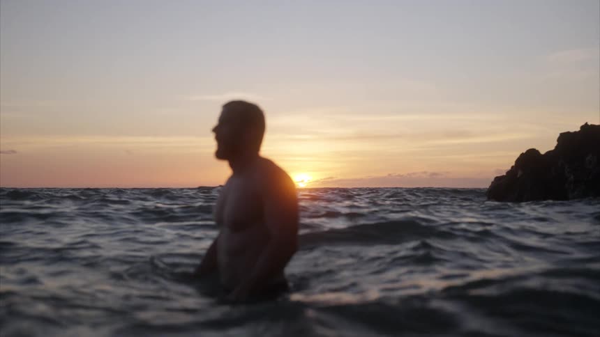 A man stands waistdeep in the ocean water enjoying the moment as ripples form from the waves with the orange setting sun in the distance