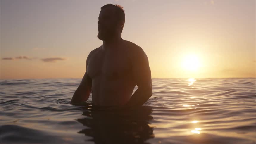 A man wades in the ocean silhouetted against a beautiful golden sunset reflecting on the water
