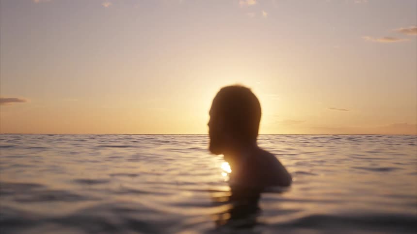 The video shows a persons silhouette as they wade into the ocean during a sunset the suns reflection shimmers on the surface of the water creating a beautiful and serene image