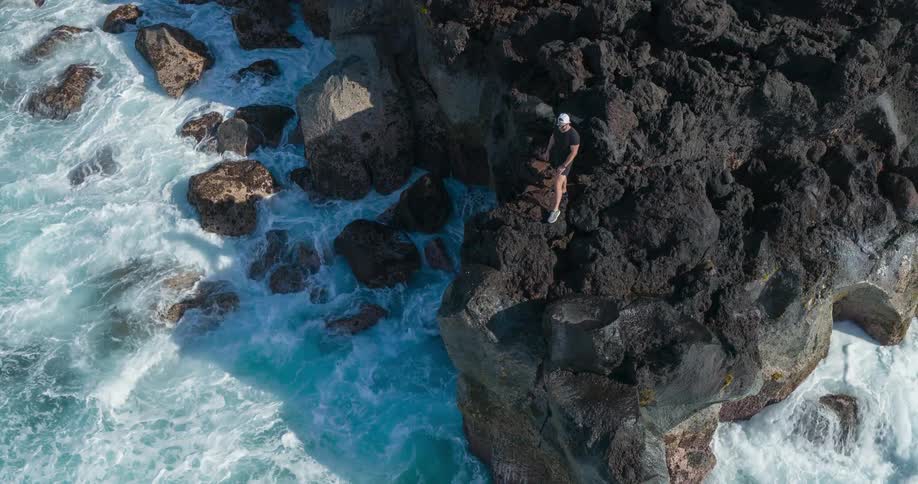 A man is standing on a rocky cliff as large turquoise ocean waves crash against the rocks below
