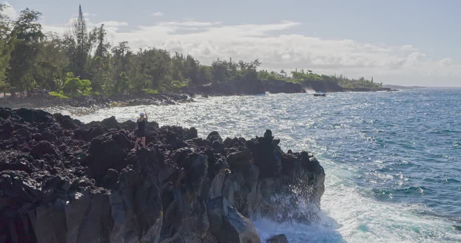 A man stands on a rocky coast watching the ocean waves smash against the rocks at midday