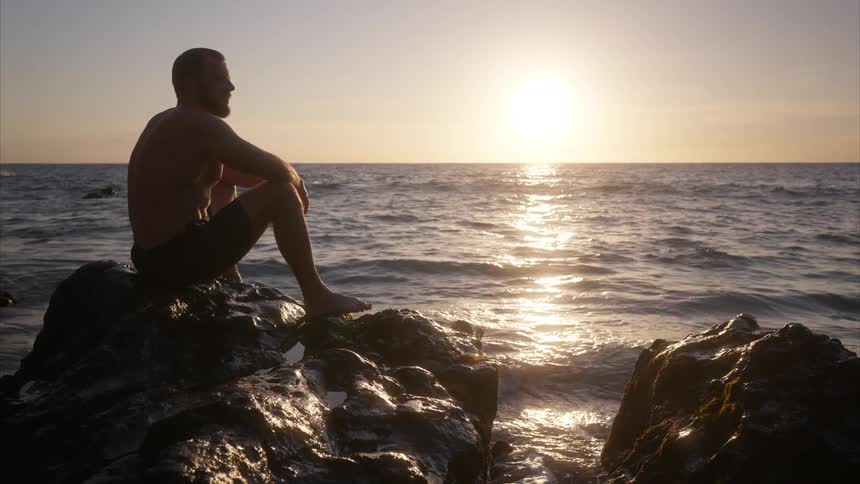 A man sits on a large rock among an ocean with gentle waves