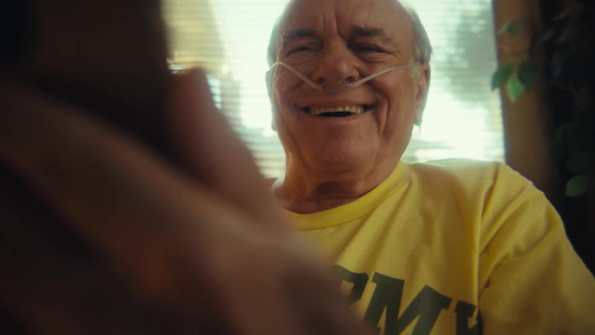 An elderly man sits in his living room wearing a yellow tshirt operating his phone and laughing while wearing a nasal cannula
