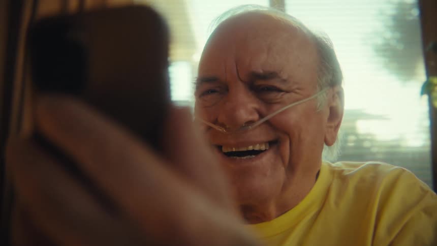 An elderly man holding his phone in front of him laughing wearing a nasal cannula in his living room at midday