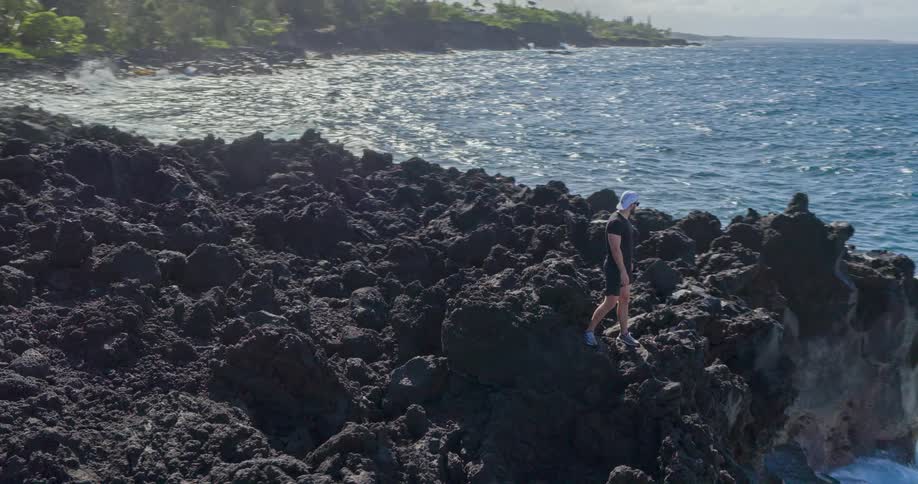 A man in sports clothes stands at the edge of a rocky cliff separating a forest from the ocean and opens his arms