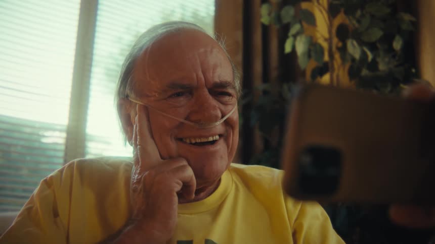 A man with white hair wearing a yellow shirt that reads us army veteran and a nasal cannula is sitting at home while smiling and touching his face with his hand as he looks down at a smartphone he holds with his other hand