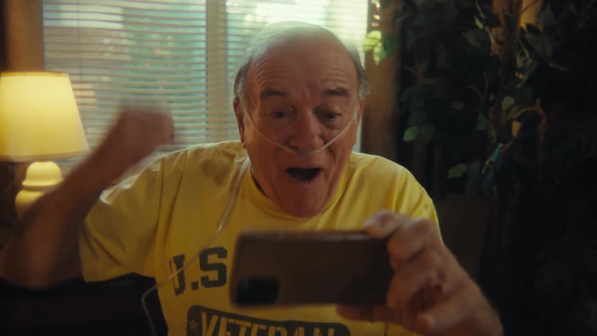 A man with white hair wearing a yellow shirt that reads us army veteran and a nasal cannula is sitting on a wheelchair smiling and laughing as he looks at a smartphone he holds in his hand
