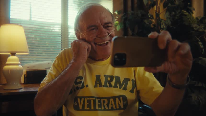A joyful man wearing a yellow us army veteran tshirt and an oxygen tube is sitting in a chair and talking on a video call on his smartphone waving and smiling at the person on the other end