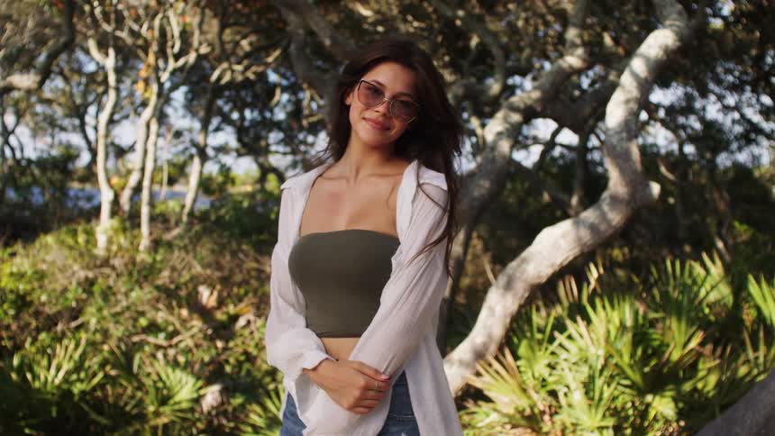 Woman wearing sunglasses smiling while standing in an environment surrounded by trees