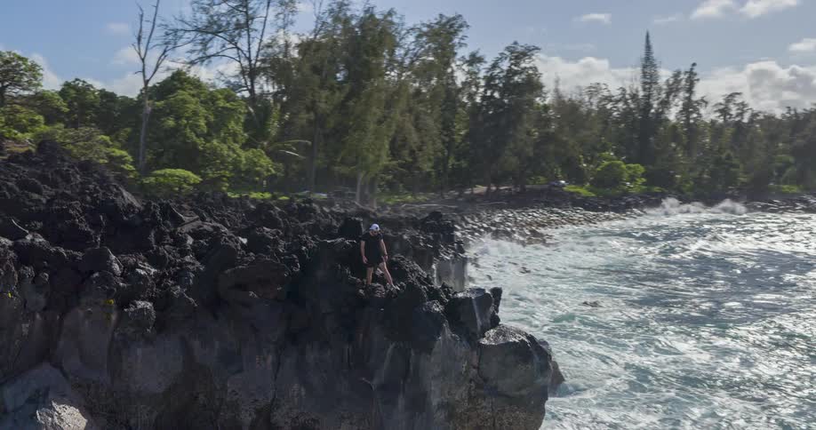 Young man stands on rocks at the edge of a cliff as waves roll in and crash against the promontory splashing high in the air