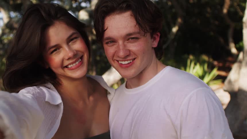 A happy young couple is smiling while taking a selfie together with the woman wearing a white shirt over a green top and the man wearing a white tshirt in a sunlit outdoor setting with trees in the background