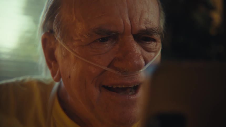 An elderly man with a breathing aid smiles as he uses a smart phone to do a video chat