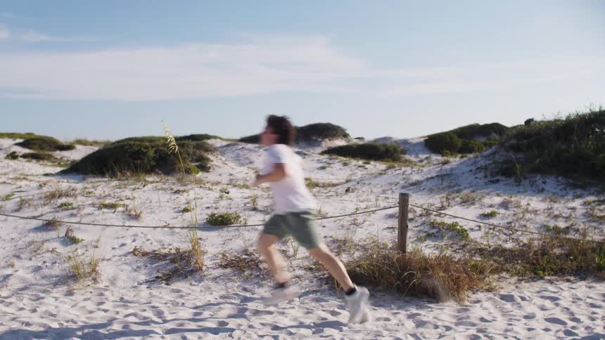 The video shows a woman running on a sandy beach shes wearing a white wrap skirt and has long wavy brown hair in the background there are sand dunes covered with some vegetation and a bright sunny sky